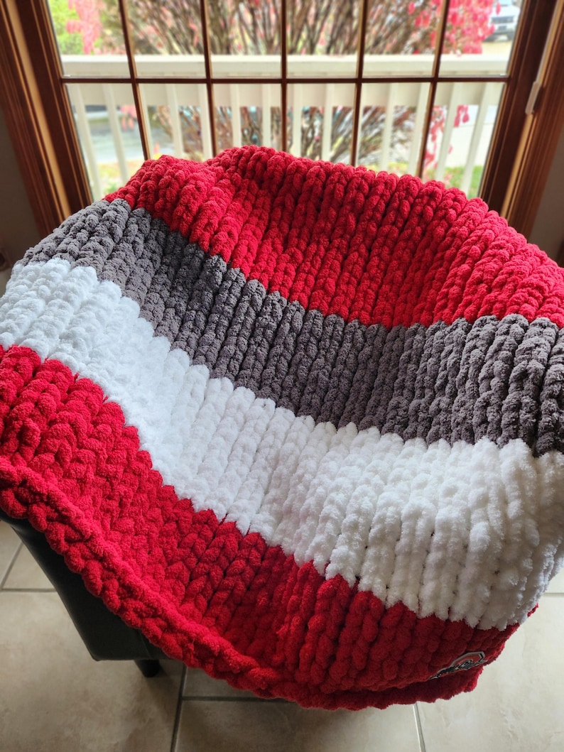 May include: A chunky knit blanket in red, white, and gray stripes. The blanket is made of a soft, textured material and is folded over a dark surface. The blanket appears to be handmade and is suitable for home decor.