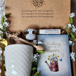 May include: A gift box containing a white mug, honey, a lip balm, a book titled "Walking with the Savior" by Max Lucado, and a gold spoon. The box is decorated with floral accents and the text "Your message here."