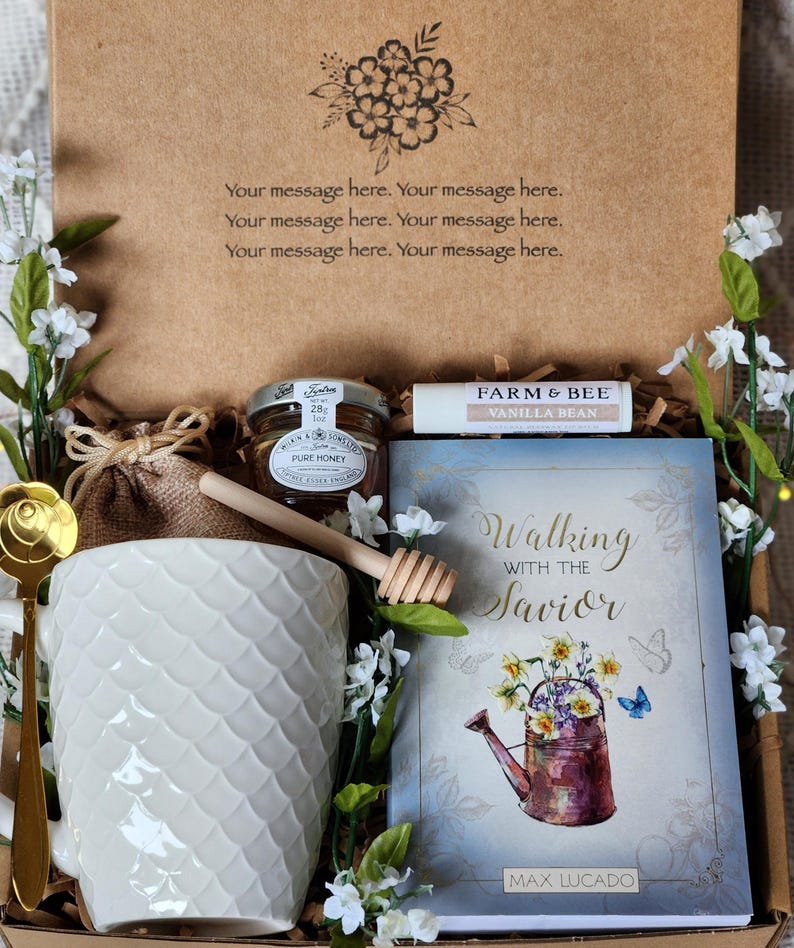 May include: A gift box containing a white mug, honey, a lip balm, a book titled "Walking with the Savior" by Max Lucado, and a gold spoon. The box is decorated with floral accents and the text "Your message here."