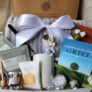 May include: A white gift basket filled with items for someone grieving. The basket includes a gray throw blanket, a white mug, a book titled "experiencing GRIEF", two boxes of tea, a jar of honey, a stick of shortbread, and a small white candle.