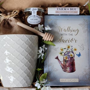 May include: A gift box with a white textured mug, a jar of honey, a lip balm, a book titled "Walking with the Savior", and a gold spoon. The book cover features a watercolor illustration of a watering can filled with flowers.