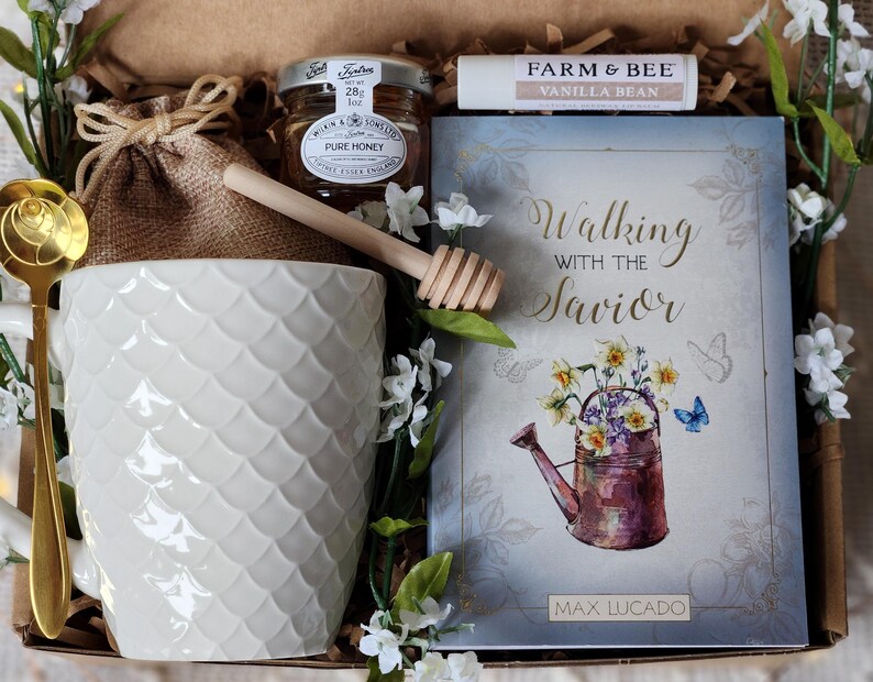 May include: A gift box with a white textured mug, a jar of honey, a lip balm, a book titled "Walking with the Savior", and a gold spoon. The book cover features a watercolor illustration of a watering can filled with flowers.