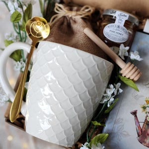 May include: A gift set featuring a white textured mug, a gold spoon, a small jar of honey, and a wooden honey dipper. The mug has a scale-like pattern. The set is decorated with white flowers and greenery, and a card.