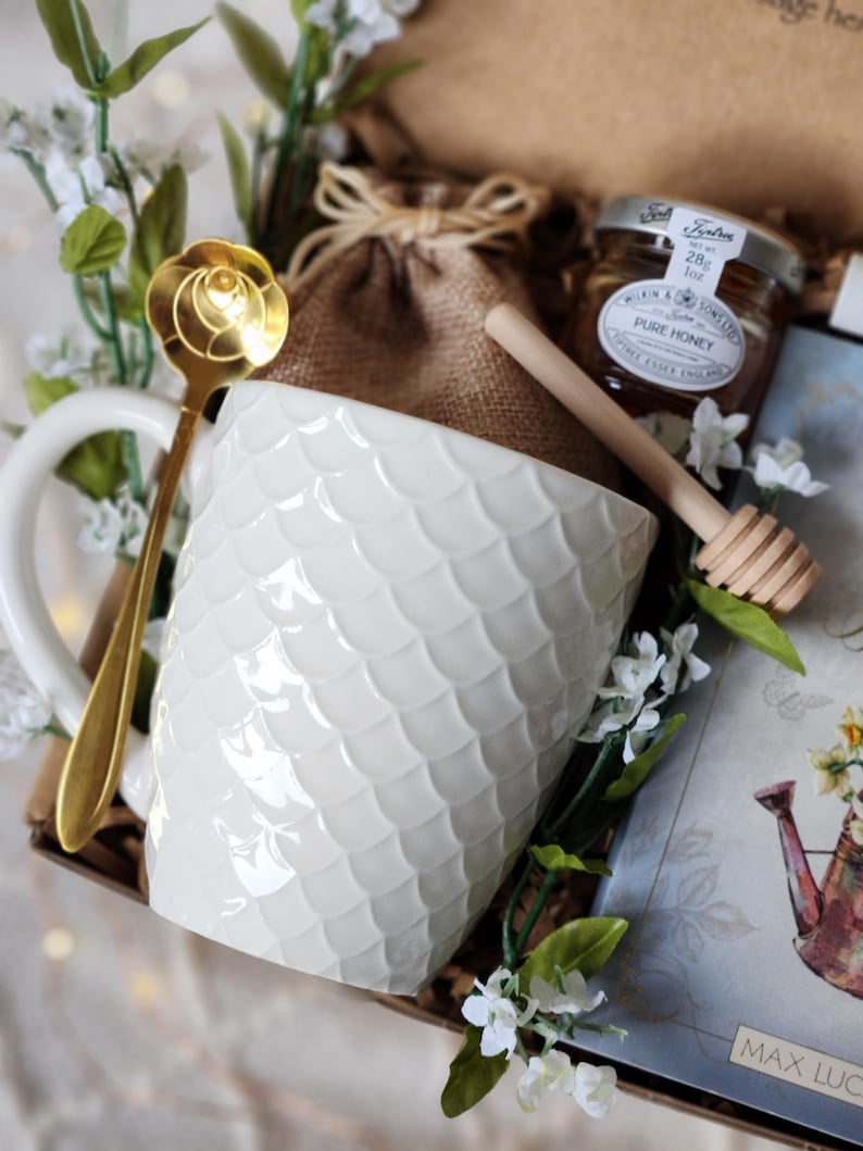 May include: A gift set featuring a white textured mug, a gold spoon, a small jar of honey, and a wooden honey dipper. The mug has a scale-like pattern. The set is decorated with white flowers and greenery, and a card.