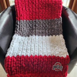 May include: A chunky knit blanket in red, gray, and white, with a red border and the Ohio State logo. The blanket is draped over a black chair, showcasing its textured design and color scheme.