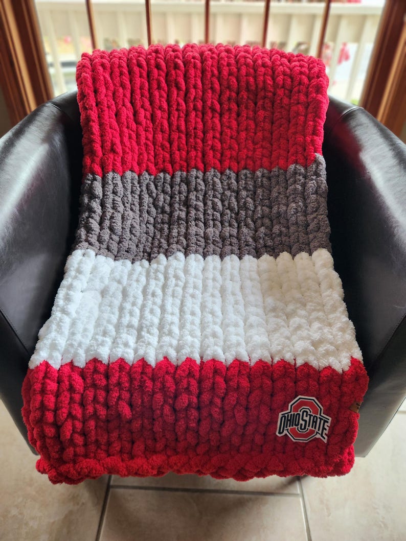 May include: A chunky knit blanket in red, gray, and white, with a red border and the Ohio State logo. The blanket is draped over a black chair, showcasing its textured design and color scheme.