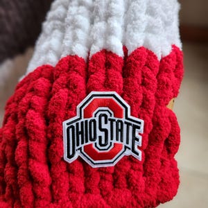 May include: A close-up of a red, white, and brown knitted item with the Ohio State logo patch. The item is made of thick, textured yarn. The Ohio State logo is in red, white, and black.