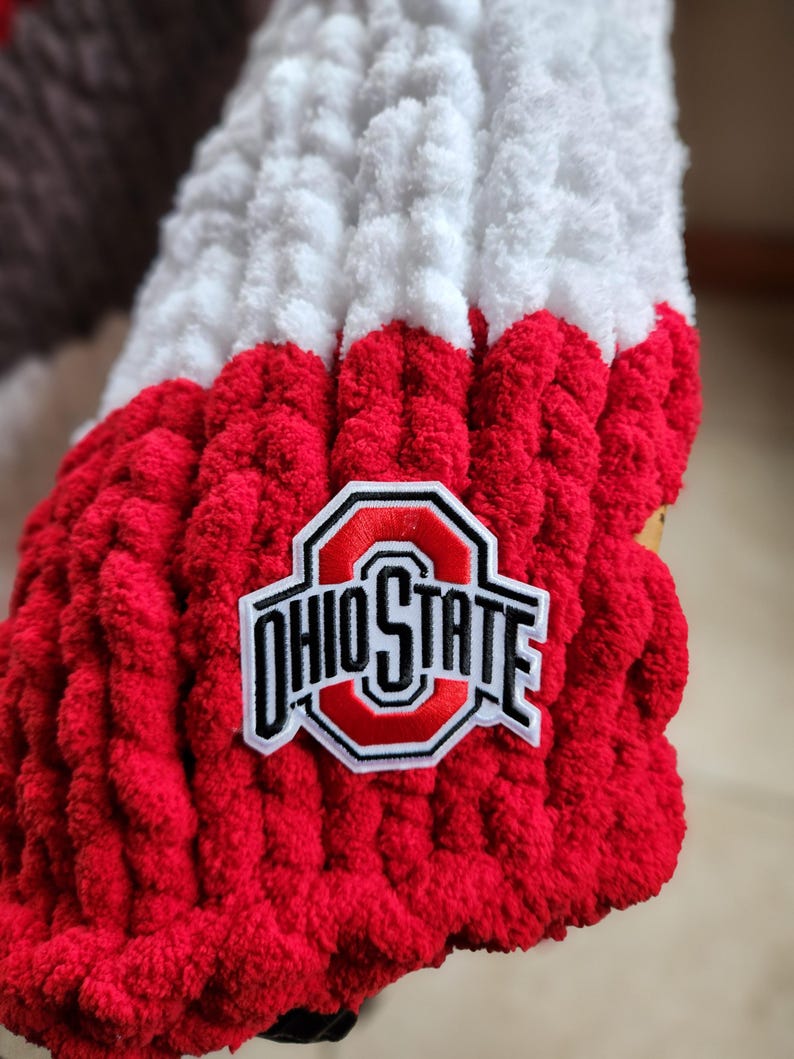May include: A close-up of a red, white, and brown knitted item with the Ohio State logo patch. The item is made of thick, textured yarn. The Ohio State logo is in red, white, and black.