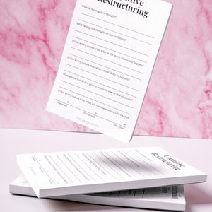 Può includere: Un blocco note bianco con il titolo "Cognitive Restructuring" e una serie di domande per aiutare con la ristrutturazione cognitiva. Il blocco note è su uno sfondo di marmo rosa.