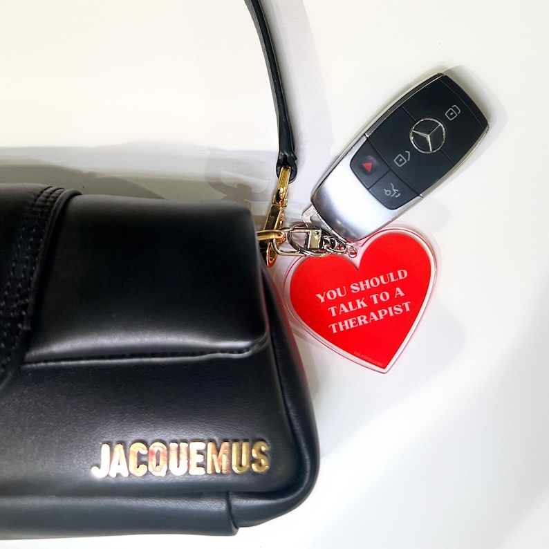 May include: A black leather handbag with the word "JACQUEMUS" in gold lettering. A red heart-shaped keychain with the text "YOU SHOULD TALK TO A THERAPIST" is attached to the bag.