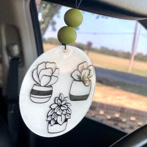 May include: A white round car charm with three black and white potted plant illustrations. The charm is hanging from a rearview mirror with green beads.