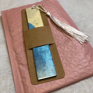 May include: Two rectangular bookmarks with blue and gold watercolor designs are displayed in a brown paper sleeve. A silver tassel is attached to the top. The bookmarks rest on a pink bubble mailer.