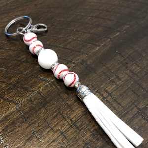 May include: White baseball-themed keychain with a white tassel. The keychain features a silver key ring and a series of white beads with red stitching, resembling baseballs.