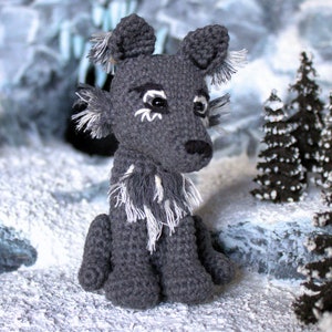 May include: A gray crocheted wolf plush toy with white fur accents sitting in a snowy landscape. The wolf has black eyes and a black nose.