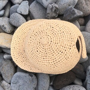 Pode incluir: Um chap&eacute;u de croch&ecirc; bege com uma aba larga e uma superf&iacute;cie texturizada. O chap&eacute;u est&aacute; sobre um leito de pedras cinzas.