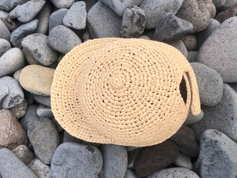 Pode incluir: Um chap&eacute;u de croch&ecirc; bege com uma aba larga e uma superf&iacute;cie texturizada. O chap&eacute;u est&aacute; sobre um leito de pedras cinzas.