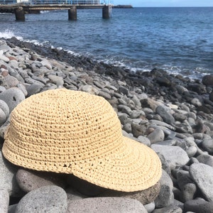 Pode incluir: Um chap&eacute;u de sol de croch&ecirc; de cor bege claro com uma aba larga. O chap&eacute;u est&aacute; sentado em um leito de pedras lisas e cinzas perto do oceano.