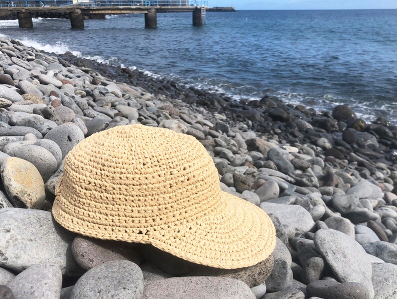 Pode incluir: Um chap&eacute;u de sol de croch&ecirc; de cor bege claro com uma aba larga. O chap&eacute;u est&aacute; sentado em um leito de pedras lisas e cinzas perto do oceano.