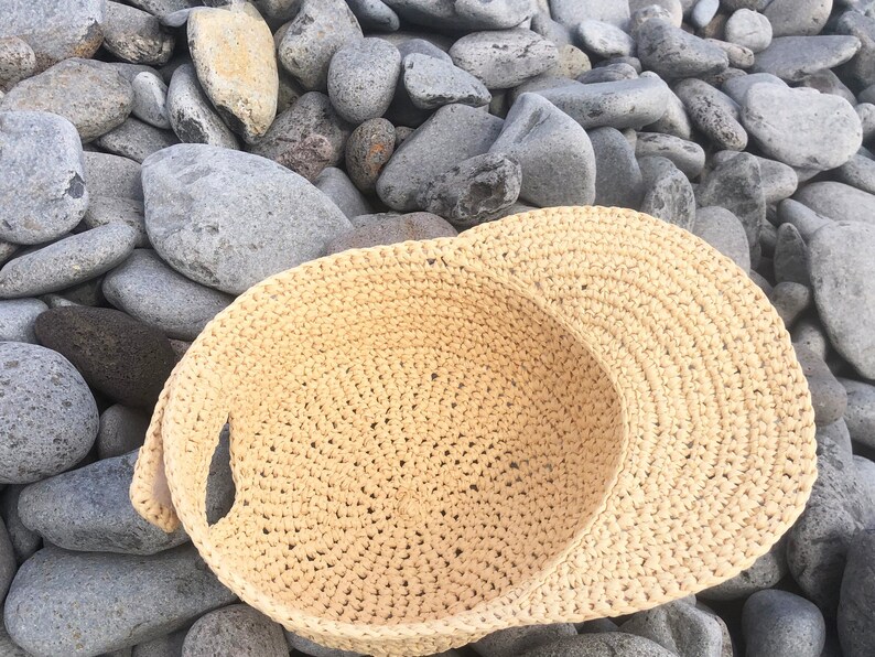 Pode incluir: Um chap&eacute;u de sol de croch&ecirc; bege com uma aba larga e uma parte superior plana. O chap&eacute;u est&aacute; sobre um leito de pedras cinzas.