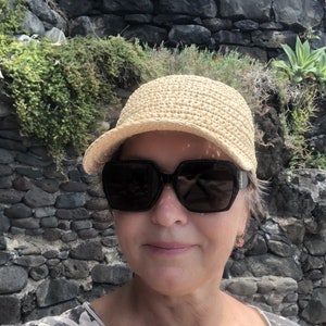 Pode incluir: Uma mulher usando um chap&eacute;u de palha de aba larga e &oacute;culos de sol escuros. O chap&eacute;u &eacute; de cor marrom claro e tem uma textura de croch&ecirc;. A mulher est&aacute; em p&eacute; em frente a uma parede de pedra.