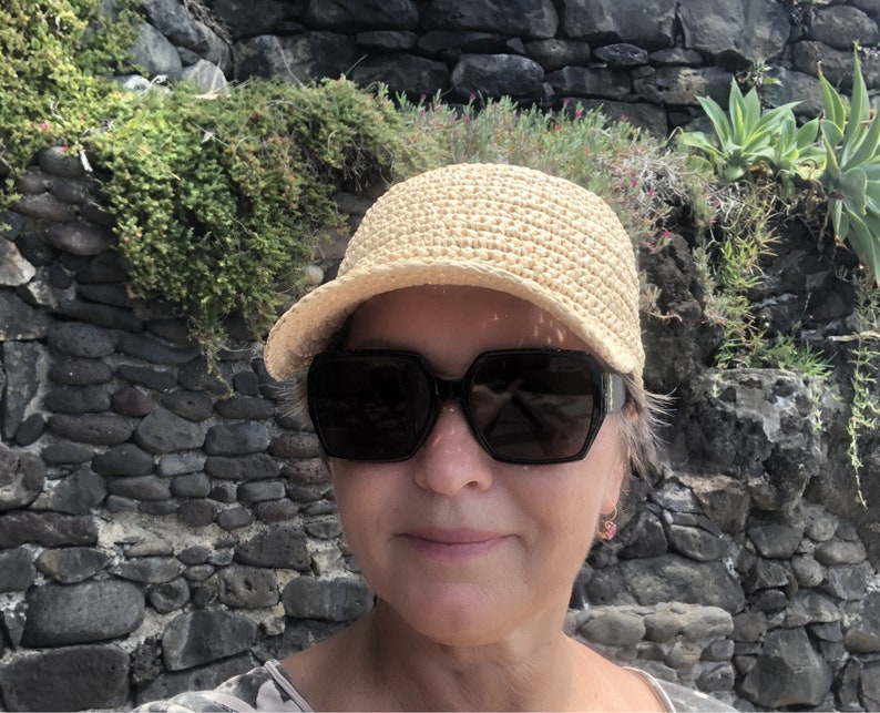 Pode incluir: Uma mulher usando um chap&eacute;u de palha de aba larga e &oacute;culos de sol escuros. O chap&eacute;u &eacute; de cor marrom claro e tem uma textura de croch&ecirc;. A mulher est&aacute; em p&eacute; em frente a uma parede de pedra.
