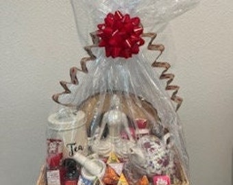 May include: A wicker basket filled with a variety of tea-themed gifts, including teacups, tea bags, a teapot, a tea infuser, a ceramic figurine, and a red bow.