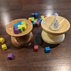 May include: Two wooden bowls with a turned base. One bowl contains an assortment of colorful, small wooden cubes. The other bowl holds a silver-wrapped chocolate candy. The bowls sit on a dark wooden surface.