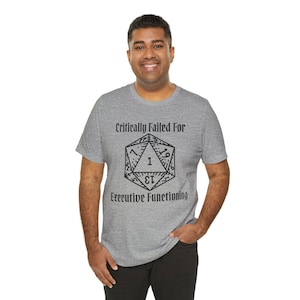 Op de afbeelding: Een grijs gemêleerd T-shirt met korte mouwen en een zwarte afbeelding van een twintigzijdige dobbelsteen met het cijfer één bovenaan. De tekst Critically Failed For Executive Functioning staat in een zwart gotisch lettertype gedrukt.