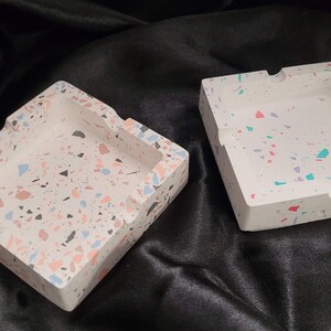 Puede incluir: Dos ceniceros cuadrados de terrazo con base blanca y motas de colores rosa, azul y verde.