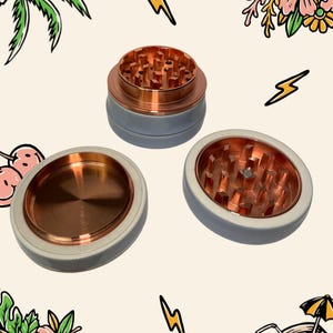 May include: A three-piece herb grinder with a matte light grey exterior and a metallic copper interior. The grinder features sharp, pyramid-shaped teeth for processing herbs, with one section showing a flat, circular copper base.