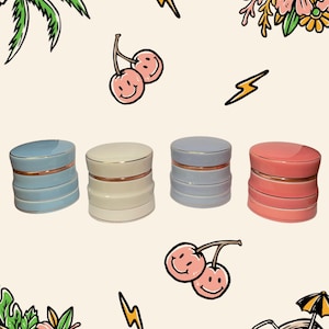 May include: Four cylindrical ceramic containers with lids, each featuring a metallic gold rim. The containers are coloured light blue, cream, light grey, and coral pink. The background is decorated with illustrations of palm leaves, cherries, lightning bolts, and a coconut drink.
