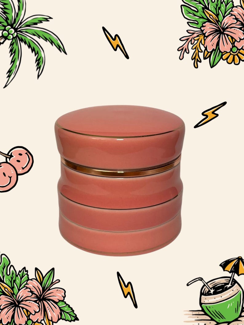 May include: A cylindrical, coral-pink ceramic container with a matching lid, featuring a smooth, glossy finish and a thin, metallic gold-coloured band accentuating the seam where the lid meets the base, set against a light cream-coloured background.