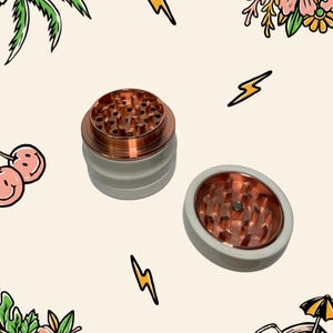 May include: A two-piece herb grinder featuring a matte white exterior and a metallic copper interior. The top section is shown upright, while the bottom section is placed flat, both revealing sharp, diamond-shaped copper teeth for grinding.