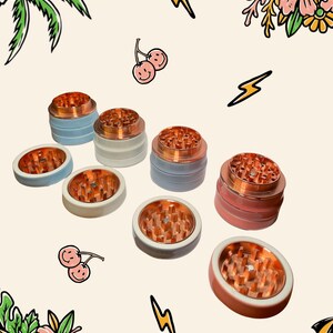 May include: A collection of herb grinders featuring metallic copper-coloured teeth and smooth, matte-finished exterior shells in shades of light blue, white, grey, and pink, arranged in a scattered pattern against a light background.