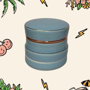 May include: A light blue ceramic jar with a cylindrical shape and a matching lid. The jar features three distinct horizontal sections separated by thin, metallic gold-coloured bands that encircle the body and the lid edge, creating a layered look.