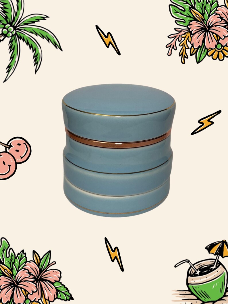 May include: A light blue ceramic jar with a cylindrical shape and a matching lid. The jar features three distinct horizontal sections separated by thin, metallic gold-coloured bands that encircle the body and the lid edge, creating a layered look.