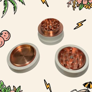 May include: A three-piece herb grinder set featuring a smooth white exterior and a metallic copper interior. The components include two pieces with sharp, pyramid-shaped grinding teeth and one flat, circular lid, all designed for manual operation.