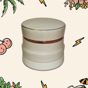 May include: A cylindrical off-white ceramic jar with a matching lid, featuring a smooth finish and a thin, metallic copper-coloured band encircling the centre where the lid meets the base. The container has a slightly indented middle section.