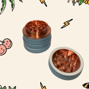 May include: A multi-part herb grinder featuring a light blue exterior and a metallic copper interior with sharp, diamond-shaped teeth. The device consists of a stacked cylindrical base and a separate lid, both showing the copper grinding mechanism.