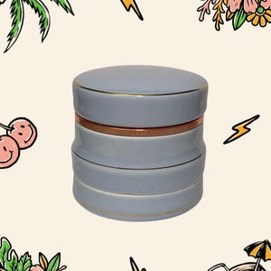 May include: A cylindrical light blue ceramic container with a lid, featuring a metallic copper-coloured band around the middle and thin gold-coloured trim along the edges of the lid and the base sections of the stackable storage jar.