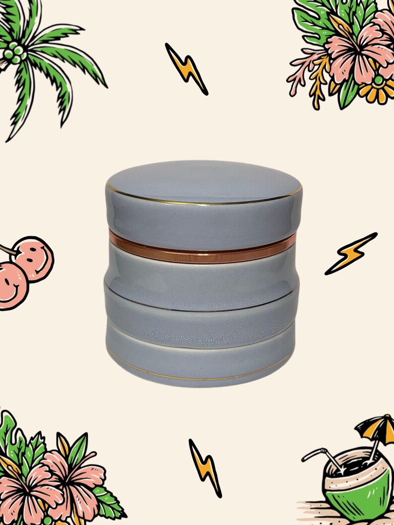 May include: A cylindrical light blue ceramic container with a lid, featuring a metallic copper-coloured band around the middle and thin gold-coloured trim along the edges of the lid and the base sections of the stackable storage jar.