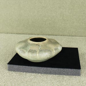 Può includere: Un vaso in ceramica dalla forma ampia e irregolare con interno scuro. Il vaso ha una smaltatura screziata verde e beige. È appoggiato su una base rettangolare nera su uno sfondo neutro. Il vaso è un oggetto decorativo.