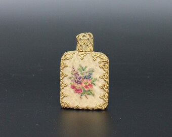 Könnte beinhalten: Eine kleine, rechteckige Parfümflasche mit einem goldenen Metallrahmen und einem gestickten Blumendesign auf der Vorderseite. Die Flasche hat einen goldenen Metallverschluss.