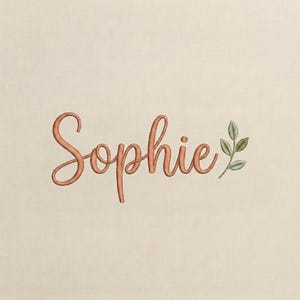 Könnte beinhalten: Gestickter Schriftzug "Sophie" in Pfirsichfarben. Ein kleiner Zweig mit grünen Blättern ergänzt den Namen auf cremefarbenem Hintergrund. Geeignet für personalisierte Artikel.