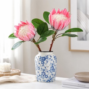 May include: Two artificial pink protea flowers with green leaves arranged in a white ceramic vase featuring a blue floral pattern. The flowers have large, layered petals surrounding a textured, cream-colored center, supported by brown stems.