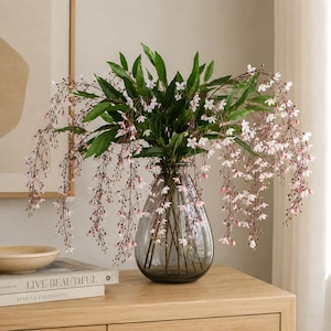May include: A clear, smoke-tinted glass vase with a teardrop shape holds a large arrangement of artificial green foliage and delicate, cascading branches featuring tiny, pale pink and white star-shaped flowers that drape over the sides.