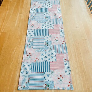 May include: A blue and pink patchwork table runner with a floral pattern and red cherries. The runner is made of cotton fabric and has a quilted design.