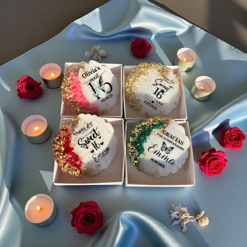 May include: Four decorative, round, white treats with colorful edges and text for a Sweet 16 celebration. Each treat is in a small white box, surrounded by red roses and lit candles on a light blue satin surface.