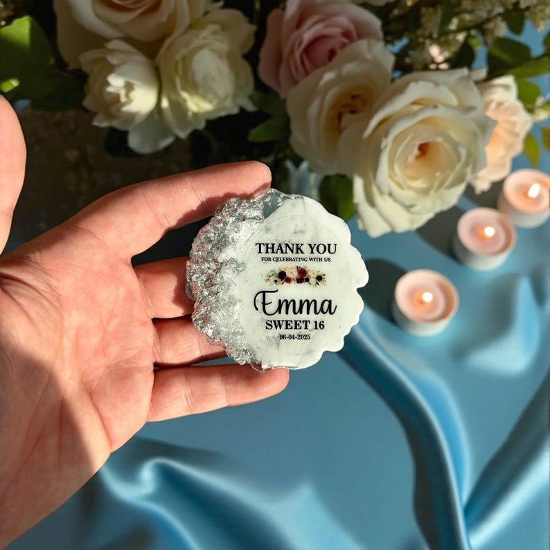 May include: A white, irregularly shaped decorative item with silver edging. The text "THANK YOU FOR CELEBRATING WITH US" is at the top, with "Emma SWEET 16 06-04-2025" below. The item is held in a hand, with flowers and candles in the background.