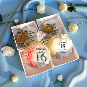 May include: Four white gift boxes with decorative cookies, gold accents, and text. The cookies have gold edges and are decorated with text such as "Sweet 16" and "Thank You". White roses are scattered around the boxes on a blue surface.